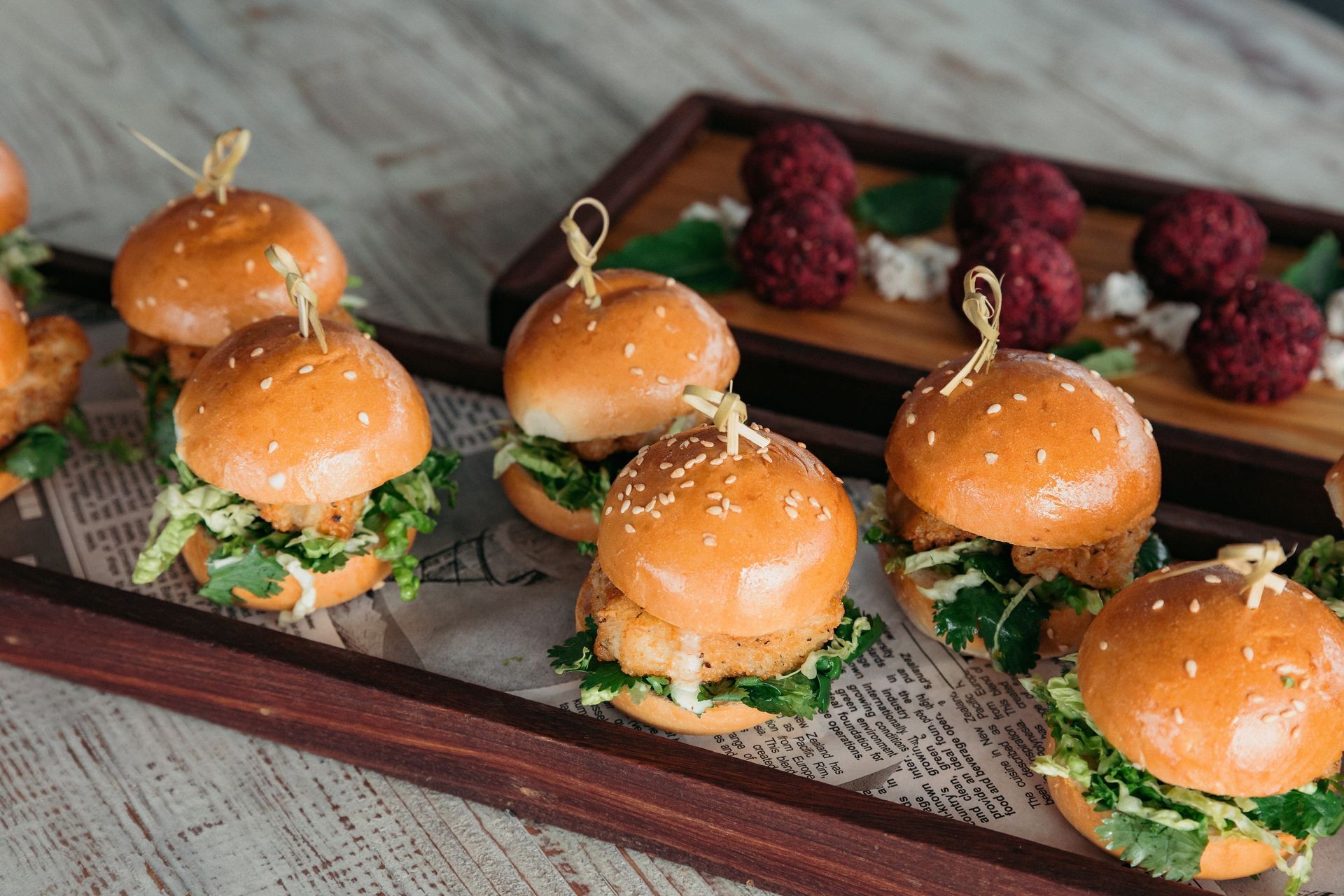 Small chicken sliders on a wooden tray with sesame seed buns and leafy greens.